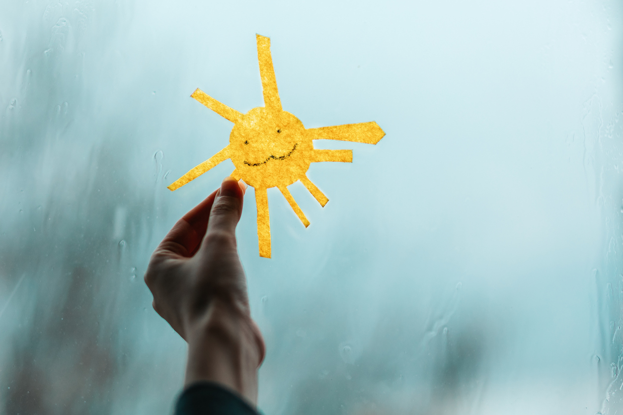 A child's hand holds the sun, cutted out of felt, against the window. Outside through the window is rain