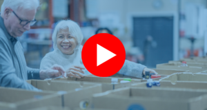 A senior gentleman and his wife work together as they pass food items between them and work to sort and pack boxes at a local Food Bank. They are both dressed casually and are smiling at one another as they enjoy their volunteer time together. |