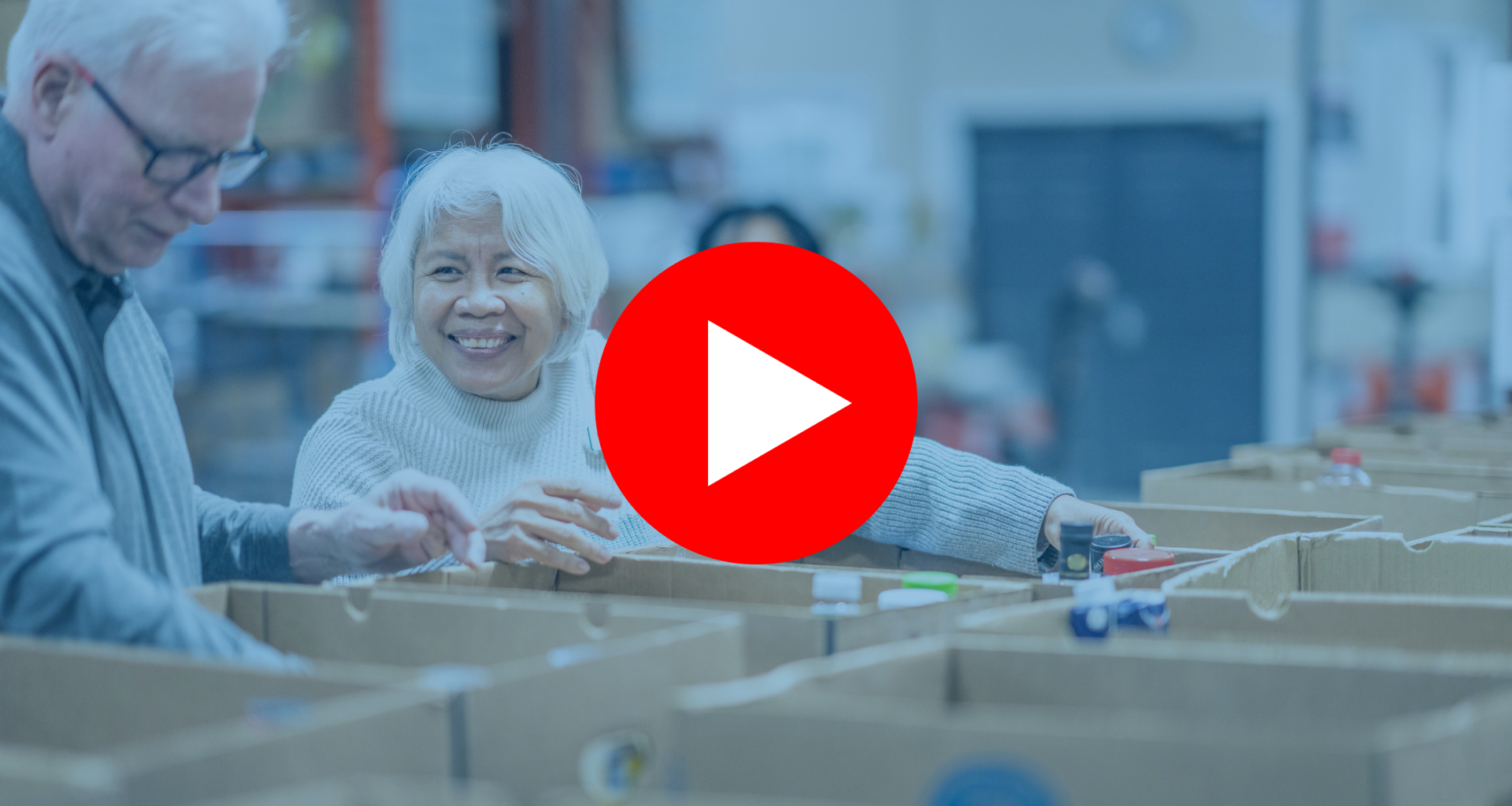 A senior gentleman and his wife work together as they pass food items between them and work to sort and pack boxes at a local Food Bank. They are both dressed casually and are smiling at one another as they enjoy their volunteer time together. |