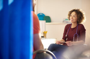A female doctor sits and chats to a male patient who we see from behind. He is partially obscured by a medical screen.