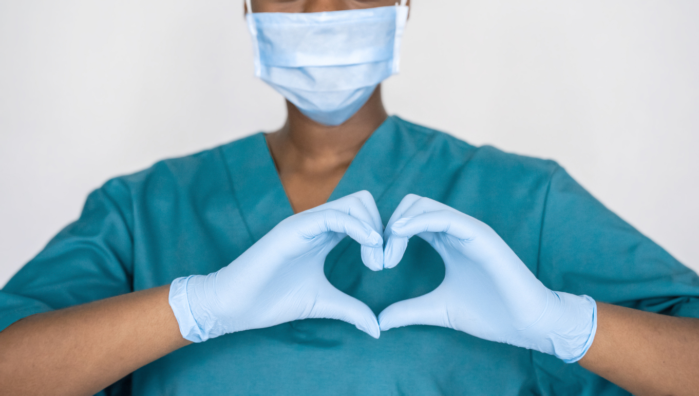 A healthcare workers wearing a mask doing a heart shape with their fingers.