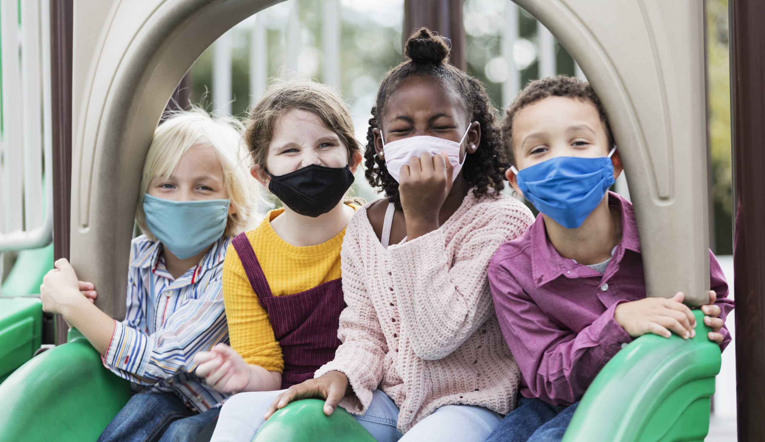 A multi-ethnic group of four children, 5 to 7 years old, playing together on a playground during school recess. They are sitting side by side on two slides, looking at the camera. They are all wearing masks, back to school during the COVID-19 pandemic, trying to prevent the spread of coronavirus.