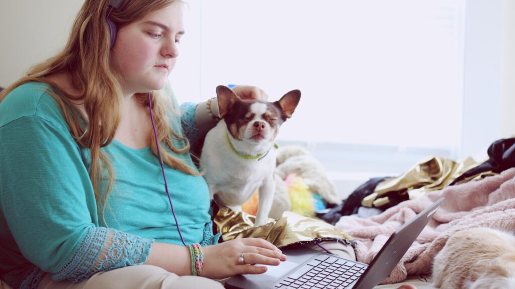 A young woman in headphones with her dog is sitting on the bed and typing on her laptop