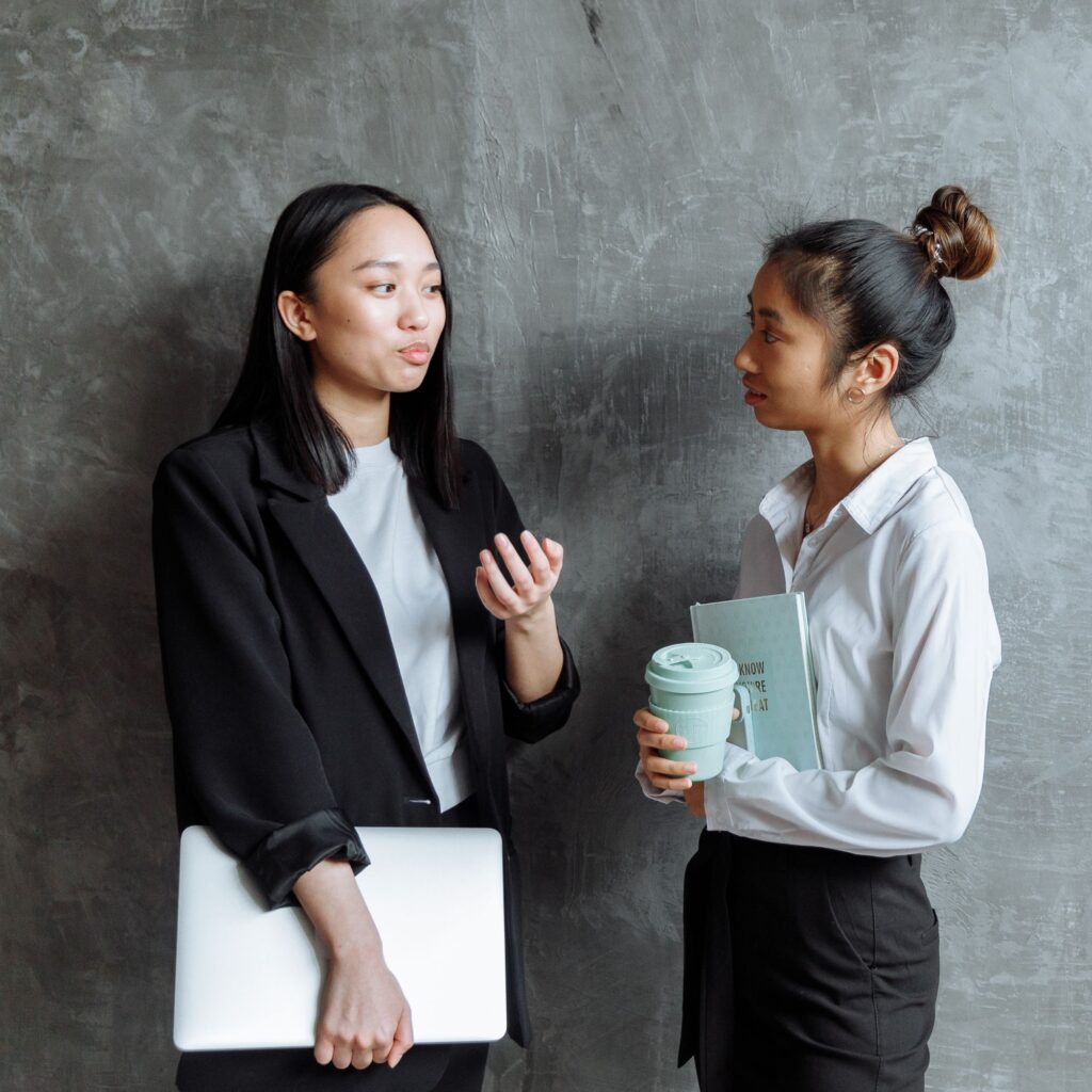 Two young women of Asian decent talking to  each other leaning on a grey cement wall