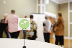 A Roots of Hope table decoration is in the foreground. In the background are blurry people standing in front of an easel.