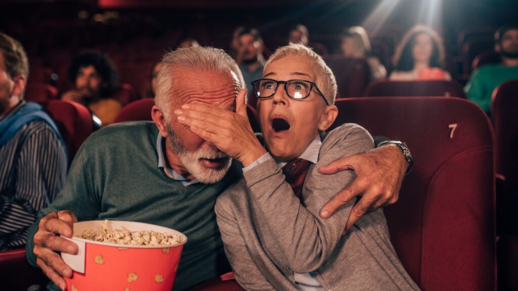 Un couple d'âge moyen dans une salle de cinéma avec un seau de pop-corn. La femme couvre les yeux de l'homme avec sa paume