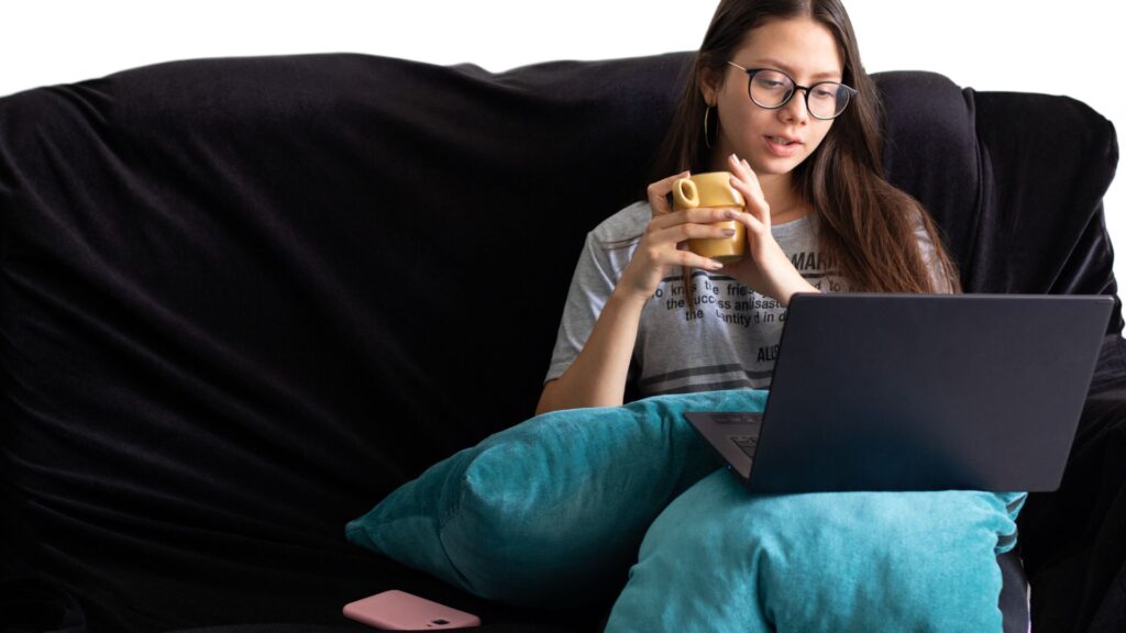 A woman sitting on a couch, using a laptop and holding a cup of coffee.