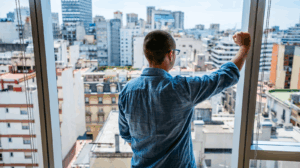 A man is looking out of an apartment window, staring at the city.